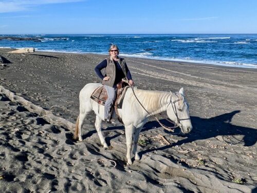 Travel writer Nancy D. Brown is riding a white horse with a western saddle and bridle. She is on Ten Mile Beach with the Pacific Ocean behind her and gray sand under her horse. She is wearing a fleece vest and sunglasses on a sunny day. 