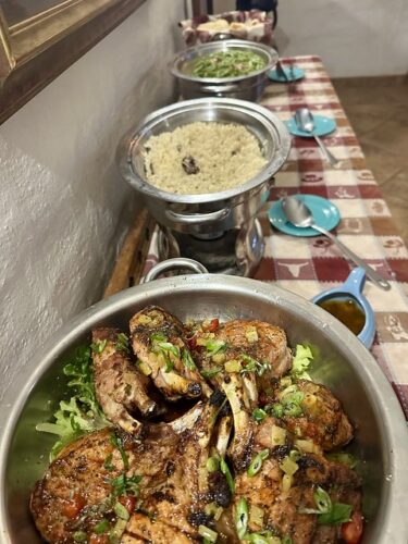 Buffet dinner, with serving dishes, on a red and white checked table cloth. First dish is a platter full of double cut pork chops. Second dish is Portobello mushroom risotto. Third dish is green beans. Last is a basket of yeast rolls with a cloth napkin under the rolls. 