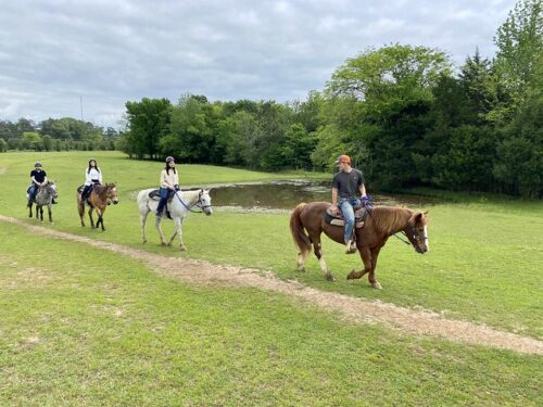 Three riders and wrangler lead the way on horseback. Horseback riding in Memphis, Tennessee with a chestnut horse and cowboy in front, white horse & rider, appaloosa horse and rider. A pond is to the right of the trail.