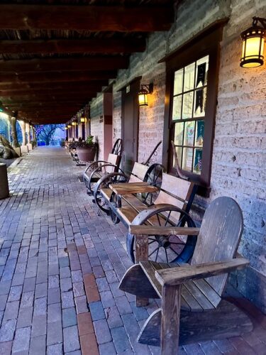 Wooden benches and rustic wooden adirondack chairs line the walkway at Tanque Verde Ranch. Brick-paved walkway holds large ceramic planter in front of one of four hotel room doors. Antique metal wagon wheels lean against the brick and adode terracotta-colored walls. 