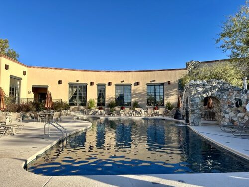 The outdoor swimming pool at Tanque Verde Ranch. Rectangular shaped pool with blue bottom and concrete patio. Backed against La Sonora Spa, the walls to the spa building show five windows into the adobe building. a rock waterfall to the right of the pool features an arch for guest to walk through. Two round patio tables with closed umbrellas are to the left of the pool. 