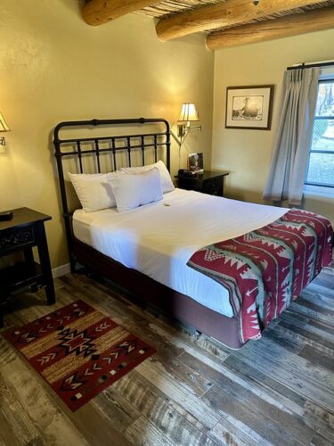 Ramada sala room with queen bed and 3 white pillows. Small wood nightstand on either side of bed. Small lamp mounted on wall, above each night stand. Wood beamed logs on ceiling with mustard-colored adobe painted walls. A large window is partially shown to right of bed. Southwestern blanket and matching area rug on wood-planked floor of hotel room. 