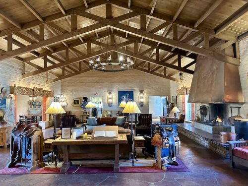 Rancho de los Caballeros lobby with high beamed wood ceiling, Wood fireplace on right. Western saddle on display on the left and right of a wooden table on display, listing daily activities. Brown leather soft is behind wood table. Four floor lamps are on the area rug covering the natural flagstone tile floor.