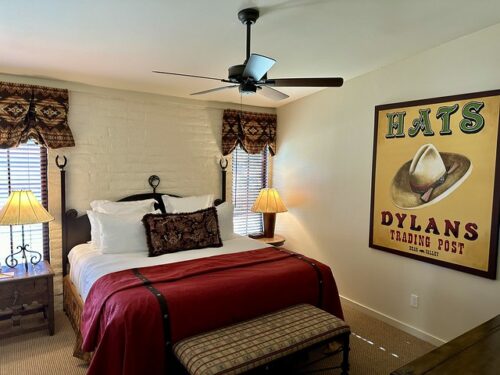 Maricopa suite bedroom with king bed, white bedding and red bedspread at the foot of the bed. Table lamps were on either side of the bed with a wood and glass-topped night stand on the left. A huge cowboy hat piece of art from Dylans Trading Post was on the right wall. 