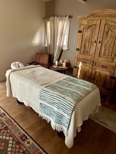 The massage table at Rancho de los Caballeros Spa. A white blanket rests over the massage table with head cradle. A wood armoire is to the right of the massage table. Cloth curtains cover a window in the spa room, to the left of the armoire. 