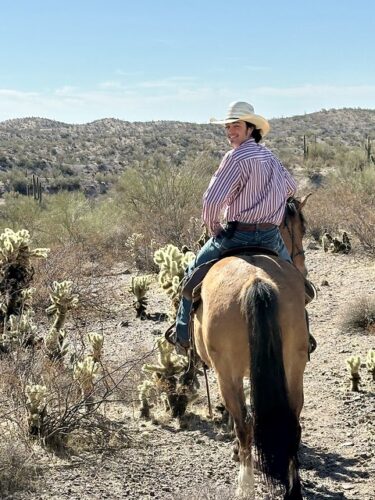 Kay El Bar Wrangler is on horseback in Arizona's Sonoran Desert. He is 23 years old, wearing a stripped shirt, jeans and a white straw cowboy hat. He has his head turned back toward the camera and his horse is pointed forward. Jumping choya cactus are on either side of the buckskin Quarter Horse. 