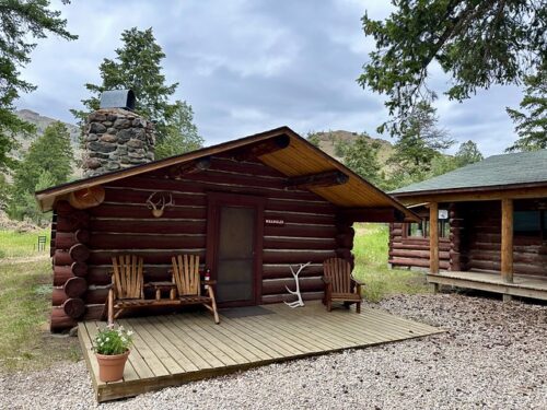 Wood log cabin with Wrangler plaque, right of front door. Wood desk with double wood bench chairs & table, all-in-one. Right of front door is wood Adirondack chair. A small potted plant of daisies is on the corner left front porch. 