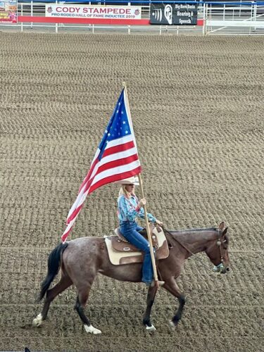 Cowgirl on horseback with blue western shirt and white straw hat. She is holding the American flag in red, white and blue colors at the rodeo. 