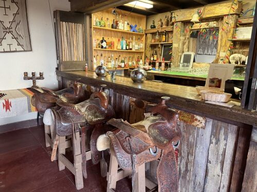 Three Western saddles serve as bar stools in the Rancho de la Osa cantina in southern Arizona. Behind the wood countertop is an honor bar lined with bottles of alcohol in various shapes and sizes.