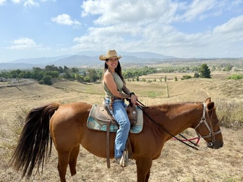 Cowgirl Maria Kurtzweil is wearing a cowboy hat, short sleeve shirt, jeans and cowboy boots. She is seated on a chestnut brown Quarter Horse and is riding in a western saddle. Behind the cowgirl is Temecula Valley landscape with dirt fields, green trees and horses in a pasture in Southern California.