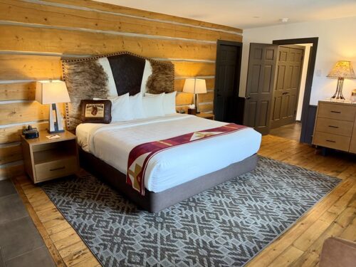 Log cabin wall, king size bed with white sheets and 4 white pillows. A headboard has cowhide of three different colors above the bed. Small wood end tables are on either side of bed with small reading lamps on the table. Geometric patterned area rug under bed with gray and silver colors. Hardwood floor within cabin. 