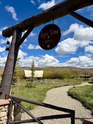 Lazy U Spa engraved on a rusted wood sawing blade. The logo sign is suspended for a heavy wood beam that frames the entrance to the gravel pathway to the spa. Green Colorado grass and 2 pine trees line the gravel rock path to the spa. A canvas covered wagon is on the left of the gravel path. Blue sky with fluffy white clouds are in the distance. 