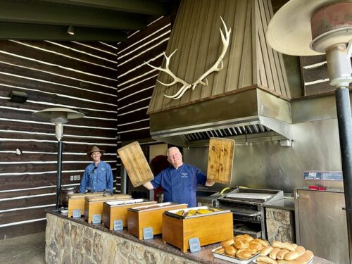 Chef wearing C Lazy U Logo blue uniform. He is holding the cover to a wood warming tray in each of his hands by the 2 of 5 warming trays. Hot dog and hamburger buns are on trays to the right of the filled buffet boxes. Behind the two servers, dressed in blue shirts is a large outdoor grill. A large elk antler rack is mounted on the outdoor stove hood. Two heat lamps are on either side of the stone-lined buffet area. 