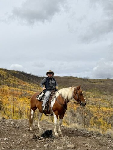 Woman, wearing cowboy hat, grey sweater and black vest with grey jeans and cowboy boots on a brown and white paint Quarter Horse with a white mane. Rider is looking at the camera, horse is looking to right. Yellow and gold Aspen trees are behind horse and rider on the dirt and rock plateau. 